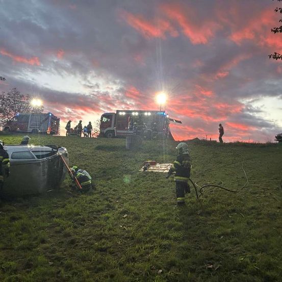 Eine Notfallszene mit einem umgekippten Auto, Feuerwehrleuten im Einsatz und Rettungsfahrzeugen auf einem Grashügel unter einem dramatischen Sonnenuntergang.