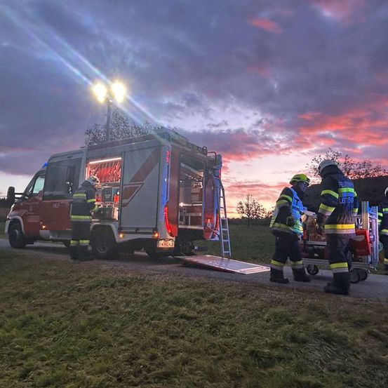 Feuerwehrleute bereiten sich darauf vor, einen Patienten auf einer Trage zu transportieren. Ein Feuerwehrwagen steht mit heruntergelassener Rampe am Straßenrand. Der Himmel ist in Orange- und Rosatöne getaucht.