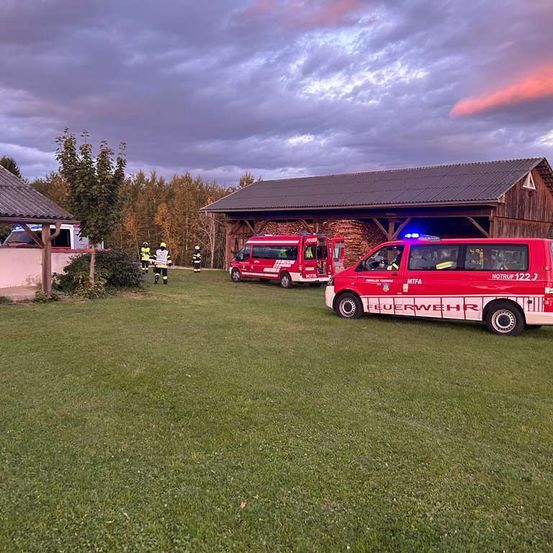 Drei rote Feuerwehrfahrzeuge und Einsatzkräfte sind auf einem Rasen vor einem Gebäude mit bewölktem Himmel geparkt.
