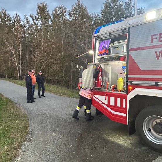 Feuerwehrleute bereiten Ausrüstung auf einem LKW vor, der auf einem asphaltierten Weg geparkt ist. Drei Personen stehen in der Nähe und beobachten. Bäume und Gras flankieren die Szene.