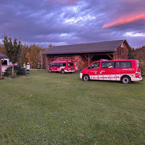 Mehrere Feuerwehrfahrzeuge sind auf einer Wiese geparkt. Dahinter steht ein großer Scheunenbau mit einem Metalldach. In der Ferne befinden sich einige Bäume und ein Haus.