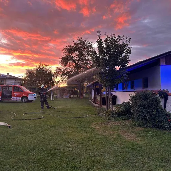Ein Feuerwehrmann spruht Wasser auf ein Haus bei Sonnenuntergang, mit einem roten Van in der Nahe geparkt.