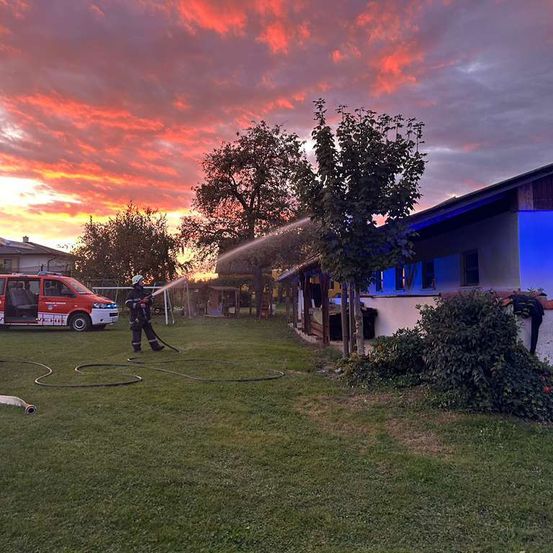 Ein Feuerwehrmann spruht Wasser auf ein Haus bei Sonnenuntergang, mit einem roten Van in der Nahe geparkt.