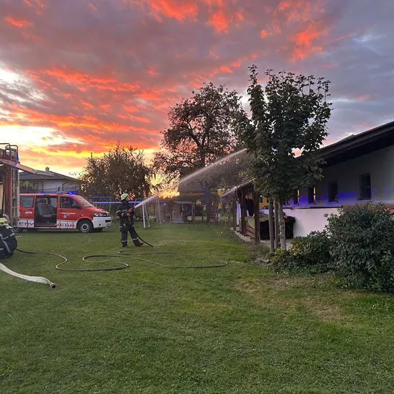 Ein Feuerwehrmann sprüht Wasser aus einem Schlauch auf ein Gebäude bei Sonnenuntergang, mit einem roten Feuerwehrwagen und geparkten Fahrzeugen in der Nähe.