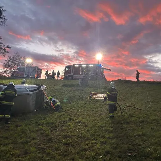Feuerwehrleute sind am Unfallort eines Fahrzeugs auf einem Hügel, unter einem rosafarbenen Sonnenuntergang.