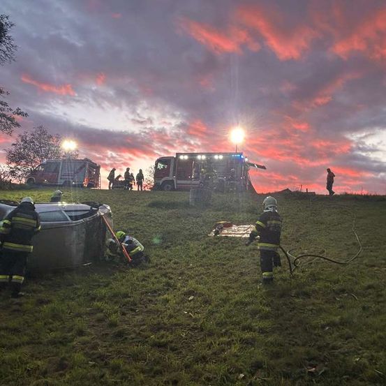 Feuerwehrleute sind am Unfallort eines Fahrzeugs auf einem Hügel, unter einem rosafarbenen Sonnenuntergang.