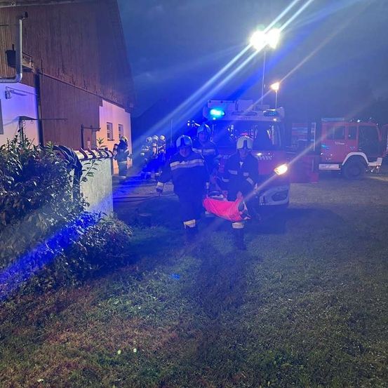 Feuerwehrleute evakuieren eine Person in einer roten Tasche aus einer Scheune bei Nacht.
