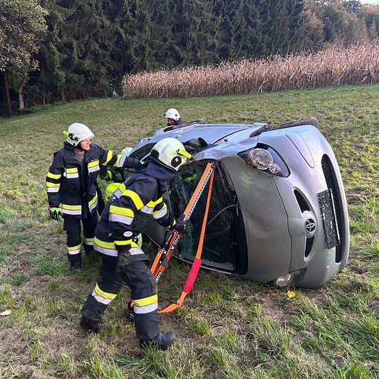 Drei Feuerwehrleute in Helmen retten ein beschädigtes Auto, das auf einer Rasenfläche auf der Seite liegt. Die Vorderseite des Autos ist stark verbeugt.
