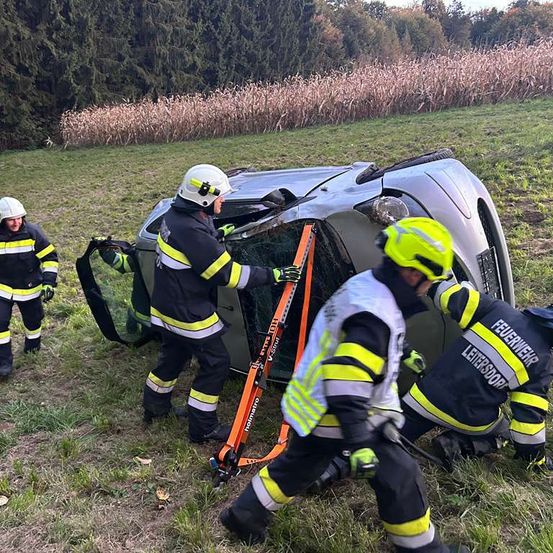 Feuerwehrleute entfernen eine Person aus einem beschädigten Fahrzeug auf einem Grasfeld. Das Fahrzeug ist umgekippt.