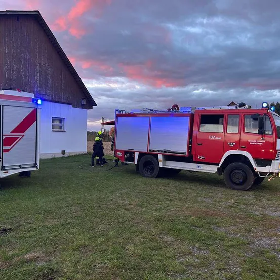 Ein roter Feuerwehrwagen ist auf einem Rasen geparkt, mit eingeschalteten Lichtern. Zwei Personen stehen dahinter und bereiten sich möglicherweise darauf vor, ein Feuer zu löschen.