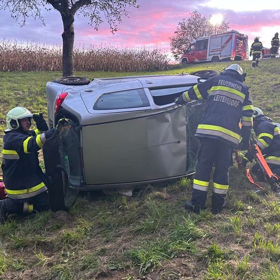 Feuerwehrleute helfen bei einem umgekippten Fahrzeug. Ein Feuerwehrmann öffnet die Tür, während ein anderer das Auto festhält. Ein Feuerwehrwagen ist oben geparkt.