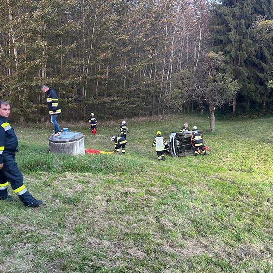 Rettungskräfte in Uniform versammeln sich um ein umgekipptes Fahrzeug auf einer Wiese. Einige kümmern sich um das Fahrzeug, während andere herumstehen.