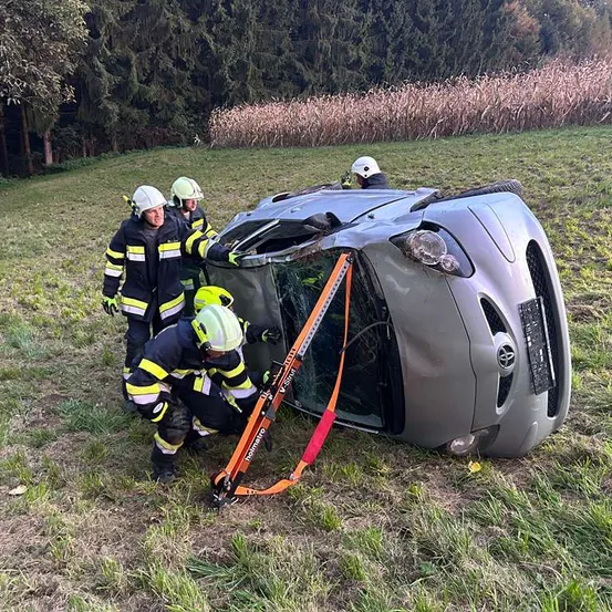 Vier Feuerwehrleute in Schutzkleidung helfen bei der Sicherung eines umgekippten Autos mit Gurten. Das Fahrzeug liegt auf der Seite mit der Front nach unten in einem Grasfeld, mit Bäumen im Hintergrund.