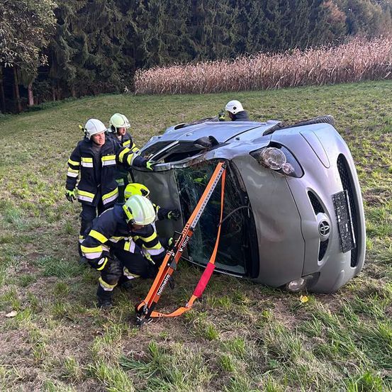Vier Feuerwehrleute in Schutzkleidung helfen bei der Sicherung eines umgekippten Autos mit Gurten. Das Fahrzeug liegt auf der Seite mit der Front nach unten in einem Grasfeld, mit Bäumen im Hintergrund.