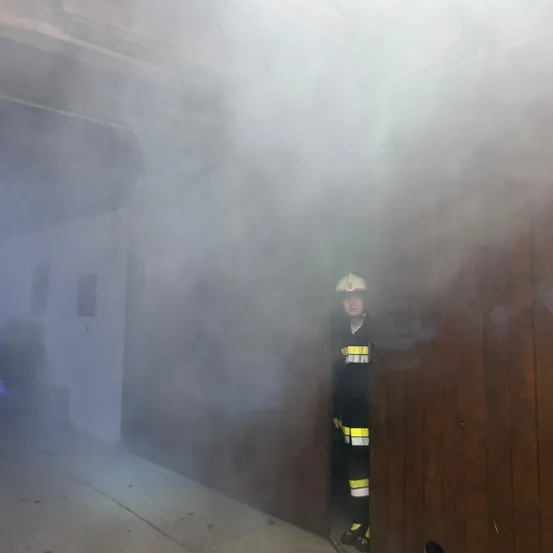 Ein Feuerwehrmann in voller Ausrüstung steht in einem raucherfüllten Raum mit einer Holztür. Der Rauch ist dick und verdeckt einen Großteil des Raumes.