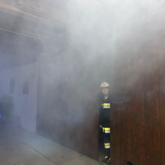 Ein Feuerwehrmann in voller Ausrüstung steht in einem raucherfüllten Raum mit einer Holztür. Der Rauch ist dick und verdeckt einen Großteil des Raumes.