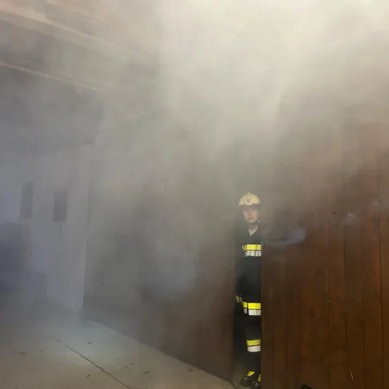Ein Feuerwehrmann steht in einem rauchgefüllten Raum, trägt einen Helm und ein gelb-schwarzes Uniform. Der Rauch verdeckt die Sicht auf den Raum.
