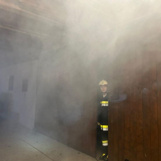 Ein Feuerwehrmann steht in einem rauchgefüllten Raum, trägt einen Helm und ein gelb-schwarzes Uniform. Der Rauch verdeckt die Sicht auf den Raum.