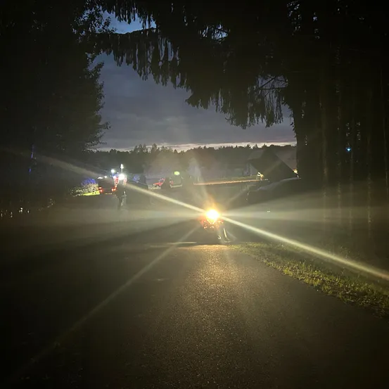 Eine Nachtszene zeigt eine Straße mit einem Motorrad im Vordergrund, dessen Scheinwerfer den Weg beleuchtet. Mehrere Fahrzeuge sind am Straßenrand geparkt, und es gibt Bäume und entfernte Lichter.