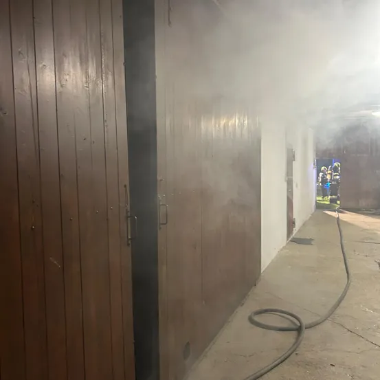 Rauch tritt aus den offenen Holztüren eines Gebäudes. Ein Schlauch liegt auf dem Boden.