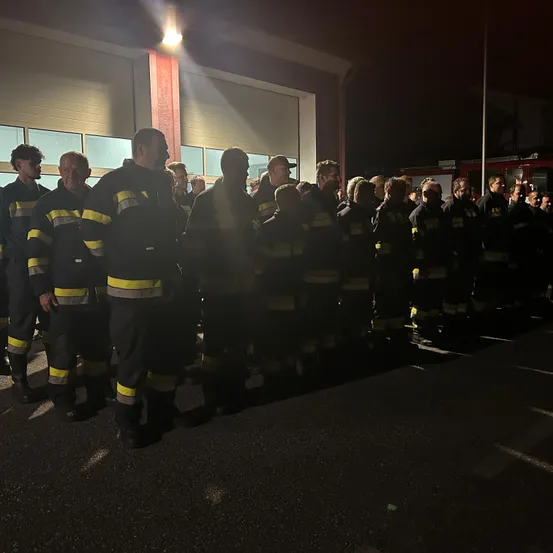 Eine Gruppe von Feuerwehrleuten steht nachts vor einem Feuerwehrhaus in Formation, beleuchtet von einem Licht.
