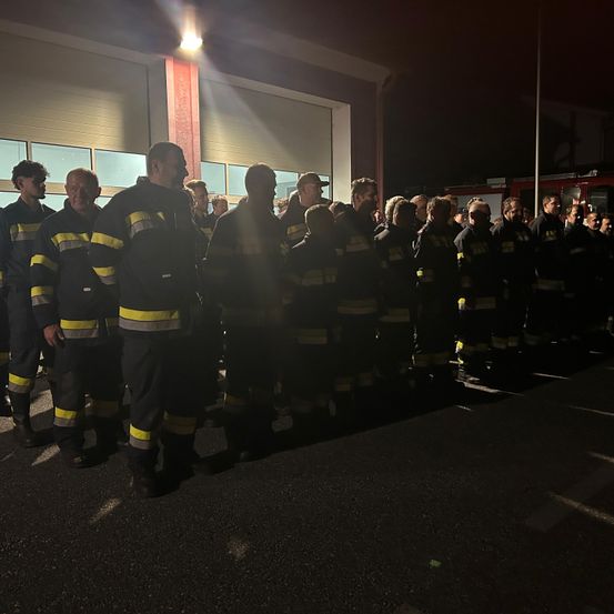 Eine Gruppe von Feuerwehrleuten steht nachts vor einem Feuerwehrhaus in Formation, beleuchtet von einem Licht.