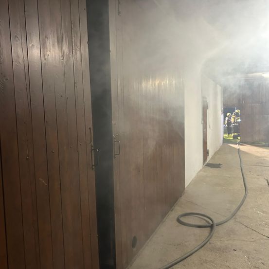 Rauch steigt aus einer offenen Holztür in einem Gebäude auf, während Feuerwehrleute draußen stehen.