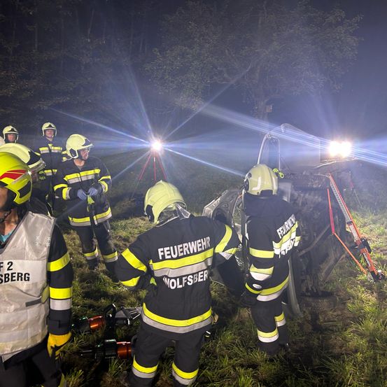 Feuerwehrleute stehen in der Nähe eines verunglückten Autos, beleuchtet von ihren Taschenlampen, in Schutzausrüstung.