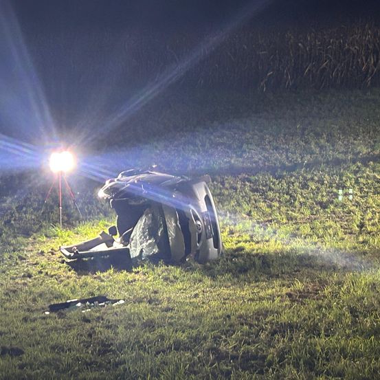 Ein Autounfall hat sich in einem Grasfeld bei Nacht ereignet. Das Fahrzeug ist umgekippt, und ein helles Licht beleuchtet die Szene.