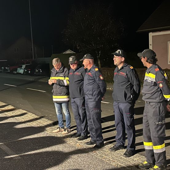 Fünf Feuerwehrleute in Uniform stehen bei Nacht an der Straßenseite. Ein Mann liest von einem Stück Papier.
