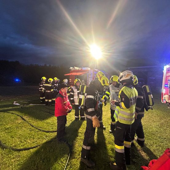 Feuerwehrleute versammeln sich auf einem Feld bei Nacht, beleuchtet von einem hellen Licht und blinkenden Rettungsfahrzeugen. Sie tragen gelbe Helme und Uniformen, einige halten Ausrüstungsgegenstände.
