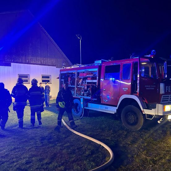 Ein roter Feuerwehrwagen steht mit blinkenden Lichtern vor einem Gebäude. Feuerwehrleute versammeln sich um den Wagen.
