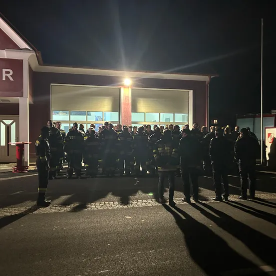 Eine Gruppe von Feuerwehrleuten steht vor einem Feuerwehrhaus bei Nacht. Das Gebäude ist von einem hellen Licht beleuchtet.