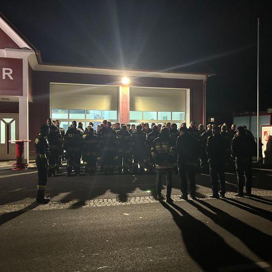 Eine Gruppe von Feuerwehrleuten steht vor einem Feuerwehrhaus bei Nacht. Das Gebäude ist von einem hellen Licht beleuchtet.