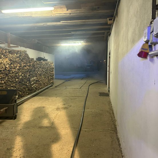 Ein Lagerraum mit Betonboden, einem Holzstapel auf der linken Seite und einem schwarzen Schlauch auf der rechten Seite. Es gibt Lampen an der Decke.