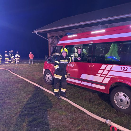 Ein Feuerwehrmann steht vor einem Feuerwehrwagen mit der Nummer 122, bei Nacht. Hinter dem Wagen ist eine Feuerwehrschläuche ausgelegt. Im Hintergrund sind andere Feuerwehrleute in der Nähe eines Holzgebäudes versammelt.