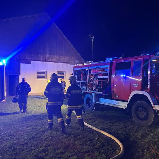 Drei Feuerwehrleute stehen bei Nacht vor einem Gebäude, einer trägt einen Helm, und ein Feuerwehrauto ist in der Nähe geparkt.