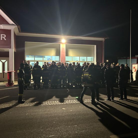 Eine Gruppe Feuerwehrleute steht bei Nacht vor einer Feuerwache, beleuchtet von einem Licht. Sie sind in voller Uniform gekleidet.