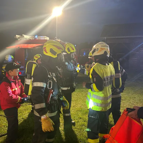 Feuerwehrleute in Schutzkleidung stehen bei Nacht auf einem Feld, mit einem Feuerwehrauto und einem Gebäude im Hintergrund.