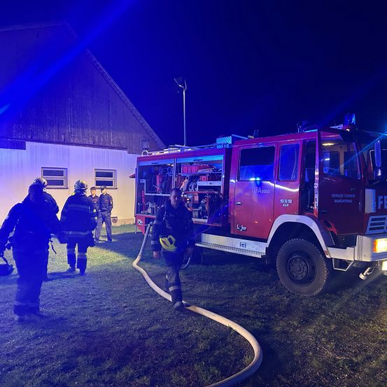 Ein Feuerwehrfahrzeug steht nachts vor einem Gebäude, Feuerwehrleute stehen in der Nähe. Ein Feuerwehrmann geht auf das Fahrzeug zu, während andere auf dem Rasen stehen.