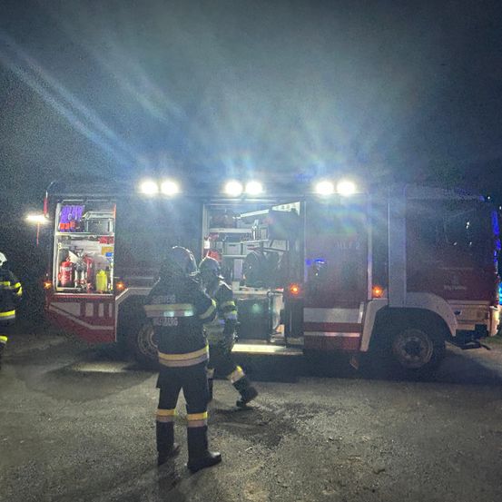 Feuerwehrleute sind an einem Ort mit einem roten Feuerwehrauto im Einsatz, das bei Nacht von Lichtern beleuchtet wird.
