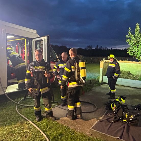 Feuerwehrleute stehen bei einem Feuerwehrauto unter einem bewölkten Nachthimmel, einige tragen Sauerstoffflaschen.