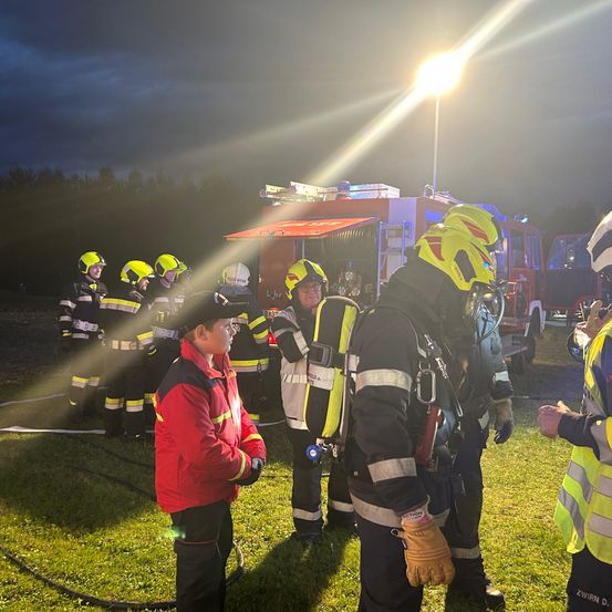 Eine Gruppe von Feuerwehrleuten in gelben und schwarzen Uniformen steht auf einem Rasen. Sie schauen einen jungen Mann in einem roten Jackett an. Hinter ihnen steht ein Feuerwehrwagen.