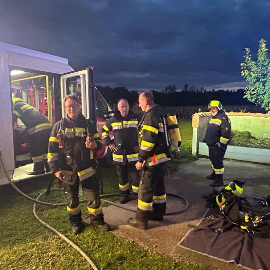 Fünf Feuerwehrleute stehen bei Nacht neben einem Feuerwehrauto. Einer lehnt sich in das Fahrzeug, ein anderer unterhält sich mit dem lehenden. Die anderen drei stehen aufrecht und beobachten.