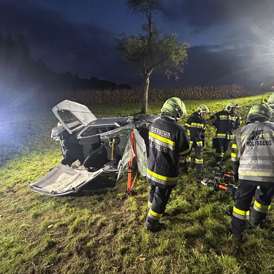 Rettungskräfte befinden sich auf einem Feld mit einem beschädigten Auto. Die Motorhaube des Autos ist geöffnet und der Kofferraum beschädigt. Die Helfer tragen Schutzausrüstung und Helme.