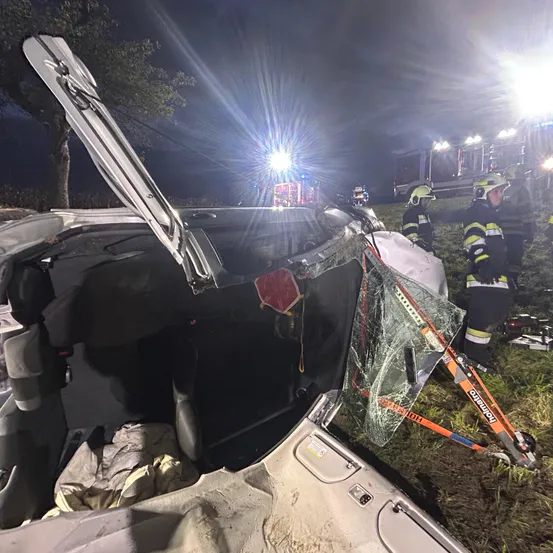 Ein Auto ist nachts am Straßenrand demoliert, mit eingeschalteten Notfalllichtern. Die Motorhaube ist geöffnet und die Windschutzscheibe ist zerbrochen. Rettungskräfte sind anwesend, von denen einer ein Werkzeug benutzt, um das Fenster zu brechen.
