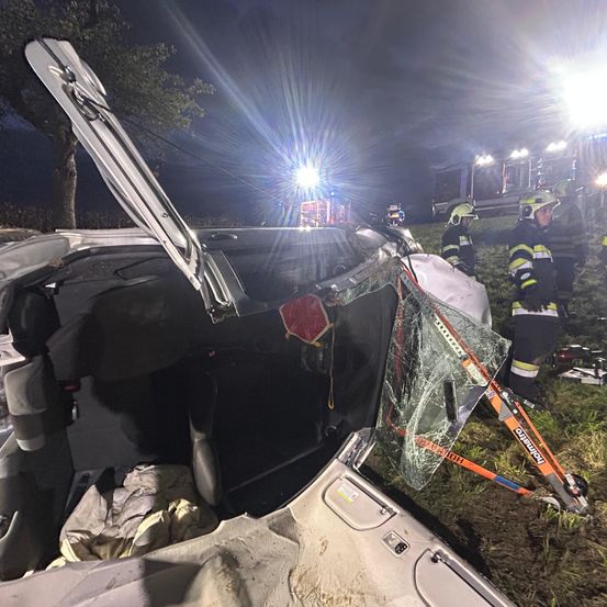 Ein Auto ist nachts am Straßenrand demoliert, mit eingeschalteten Notfalllichtern. Die Motorhaube ist geöffnet und die Windschutzscheibe ist zerbrochen. Rettungskräfte sind anwesend, von denen einer ein Werkzeug benutzt, um das Fenster zu brechen.