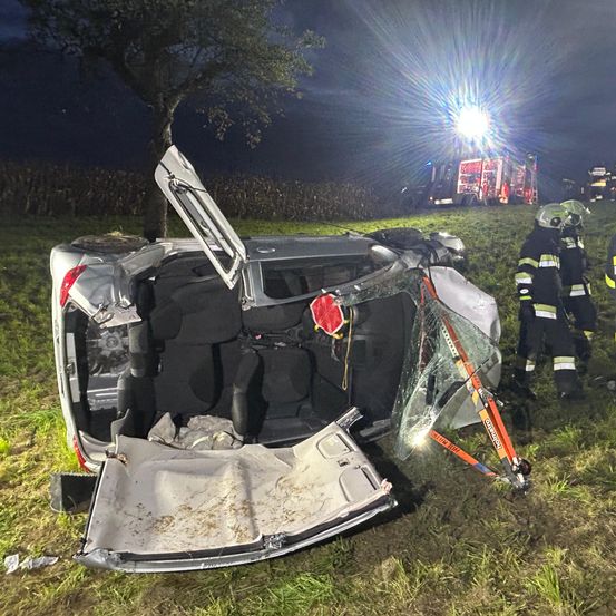 Ein umgestürztes Auto mit offenen Türen liegt auf einem Grasfeld. Rettungsdienste sind anwesend, mit einem Feuerwehrauto und Personal in der Nähe.