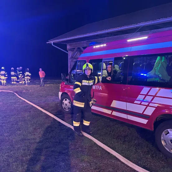 Ein Feuerwehrmann steht neben einem roten Feuerwehrwagen mit der Aufschrift MTFA. Dahinter stehen mehrere andere Feuerwehrleute.
