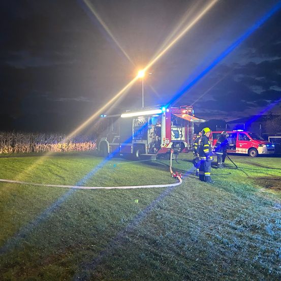 In der Nacht stehen Feuerwehrleute auf einem Rasenfeld mit einem Feuerwehrwagen und einem Krankenwagen. Sie benutzen einen Schlauch. Der Himmel ist dunkel.